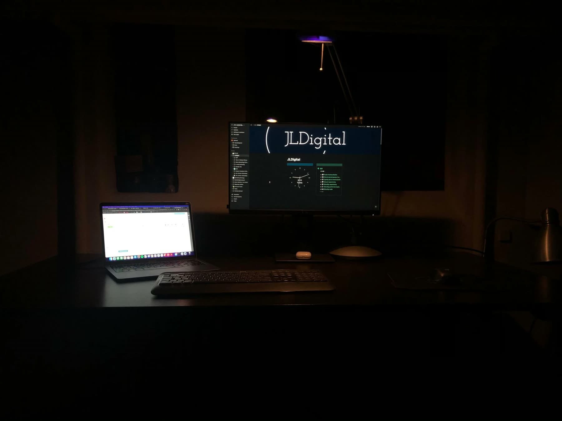 Et tidligt JLDigital-arbejdssetup med laptop og skærm i et mørkt arbejdsrum